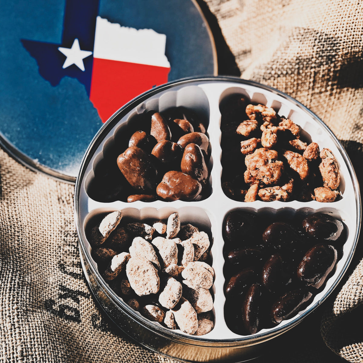 A tin with compartments containing various flavored pecans, placed on a burlap fabric with a Texas state flag design in the background.